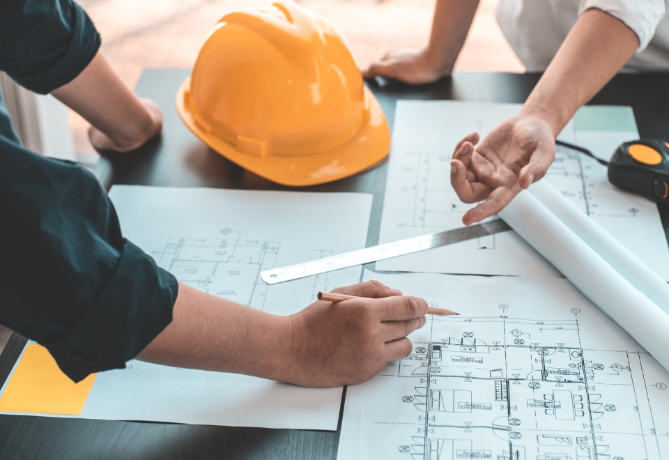 Hands reviewing building plans with a hard hat on the table, symbolising the importance of owner builder insurance.