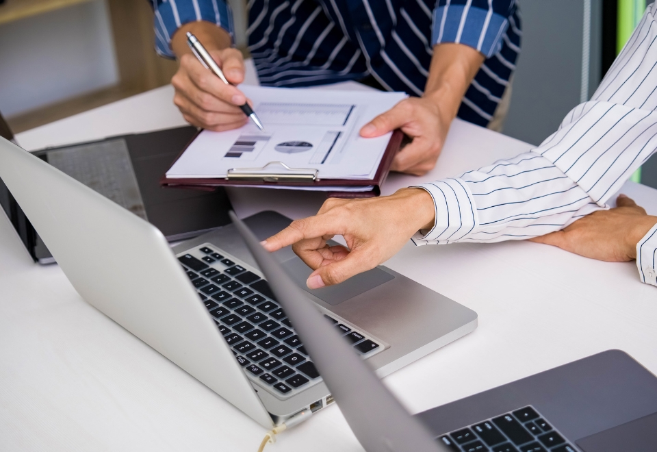 Two professionals reviewing charts on a laptop and clipboard, representing the importance of business insurance for startups.