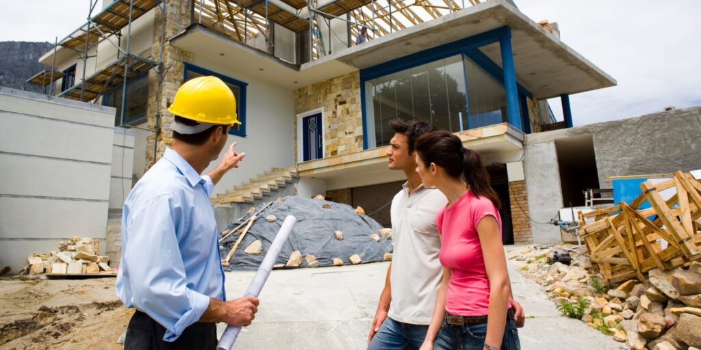 A contractor in a yellow hard hat discusses a building project with two clients in front of a partially constructed house.